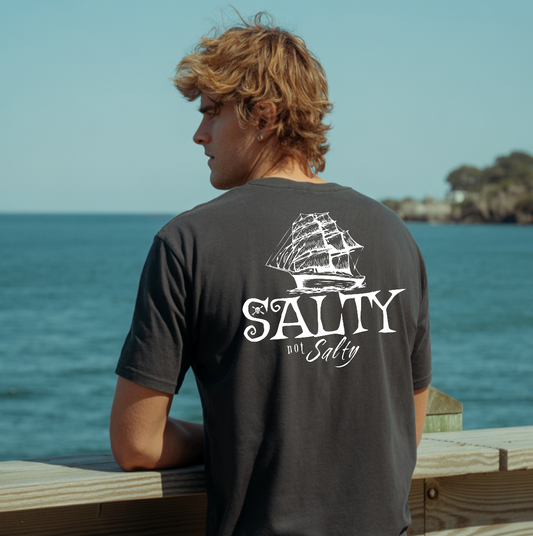 Person wearing a 'Salty' t-shirt with a ship graphic by the ocean.